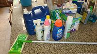 Photo showing overall lot contents including Swiffer mop, boxed cat litter, multiple bottles of laundry detergent and cleaning sprays arranged on a counter.