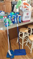 Cleaning supplies displayed on kitchen countertop including cleaning liquids, brooms, dustpan, duster, mop, and bucket.
