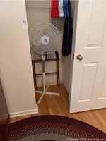 View showing the braided rug on the floor with part of the white floor fan and wooden luggage rack behind it along a wall.
