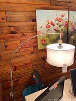 Brushed stainless steel arc floor lamp with white fabric drum shade in an interior with wooden wall and flower painting.