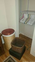 Tall wicker hamper with reddish lid along with two wicker baskets and metal shoe rack with plastic hangers on shelf above.
