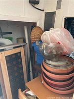 Photo showing a stack of various terracotta pots, a clear textured bowl on top, and a wicker basket in the background along with other miscellaneous garden items.