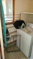 Laundry supplies arranged in laundry room including wooden and metal drying racks, ironing boards, iron on top of dryer, and collapsible laundry basket.