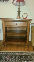 Front view of one wooden nightstand showing drawer, two shelves, and rounded bun feet. Lamp and clock on top for scale.