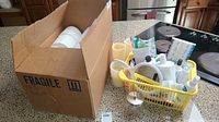 Cardboard box labeled 'Fragile' filled with multiple rolls of white toilet paper next to a plastic basket with various personal care bottles and flameless candles on a counter