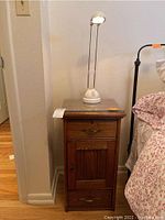 Small oak cabinet with glass top, two drawers and a door, with a white adjustable desk lamp on top, placed next to a bed.