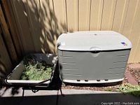 Rubbermaid outdoor storage box next to garden cart with plant material inside both items outdoors by a wooden fence.