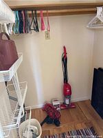 Red upright Dirt Devil vacuum with cord, handheld Dirt Devil vacuum, plastic hangers on rod, and black boot trays on closet floor