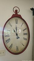 Large oval wall clock with red frame and Roman numeral face hanging on wall