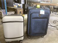 Two carry on suitcases, hard shell white/light gray and soft fabric dark blue, standing upright side by side on concrete floor.