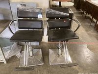 Front view of two black salon chairs with chrome bases and armrests, showing shapes and conditions.