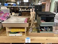 Shelf view of art supplies lot showing wooden mannequin figures, pencil crayons in wooden box, and art supplies in storage boxes.