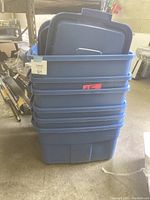 Stack of six blue Rubbermaid storage bins with lids seen on top. The bins are stacked inside each other, showing their size and color. The bins are placed in a warehouse or storage area.