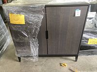 Front view of the wooden cabinet wrapped partially with plastic film, showing the two doors and legs.