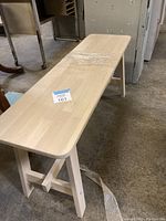 Full view of the light wood bench showing top and legs, taped center area, placed on a concrete floor.