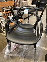 Front angle view of black plastic chair showing distinctive open arm and back design and integrated legs wrapped in protective plastic.