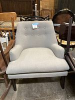 Front view of the grey thick cushioned armchair showing the whole chair including legs and armrests, placed among other chairs.