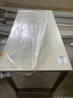 Full view of white rectangular desk with black legs, plastic wrapped on top.