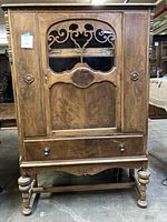 Front view of wood jewelry stand featuring an arched door with decorative cutout and peek a boo window, one large drawer at the bottom, and turned legs.