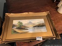 Framed watercolour landscape painting depicting lake, mountains, trees, and a house in a decorative gold frame with visible broken corners.