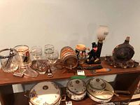 Full view of the bar lot items arranged on table including glassware, oil lamp, stein, bar tools, wooden barrel and ceramic cookware.
