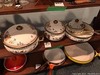 Close-up of decorative ceramic cookware with floral design and brass handles with lids on middle shelf, plus some metal pans on bottom shelf