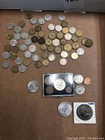 Overview photo showing a variety of silver, gold-tone and bronze coins spread on cardboard with a 1982 Queen Elizabeth commemorative coin in protective case, some additional coins outside the case.