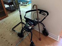 Lot of health equipment including a four-wheeled walker with seat and brakes, a wooden cane, and a digital blood pressure monitor.
