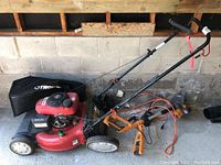 Overall view of red Troy-Bilt mower, grass bag attached, and orange Worx trimmer laid on concrete floor