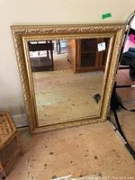 Full front view of the large gold framed mirror resting against a wooden floor and wall showing its overall dimensions and style.