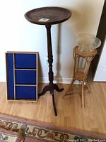 Tall wooden pedestal plant stand with round top, small wicker plant stand, and a painting leaning against the wall.