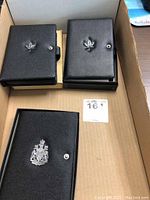 Three black leatherette cases with maple leaf and coat of arms emblems on their covers inside a cardboard box.