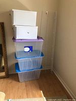 Stack of three large clear plastic totes with blue lids and two smaller white storage bins on top, situated on a wooden floor.