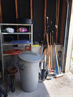 Overall view of lot showing metal shelving unit with garden supplies on shelves, garbage can with lid, women's rubber boots beside it, and long handle yard tools against the wall.