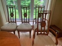 Four wooden dining chairs, two with armrests and two without, arranged near window. Beige upholstery on seats, carved wooden backs with oval medallion. Light marks visible on some seats.