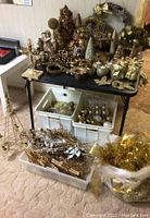 Wide view showing gold-themed Christmas decor assortment on black folding table with several plastic tubs of additional decorations below and in front of the table.