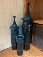 Three blue textured glass vases with tall pointed lids arranged in a corner by wall and wooden furniture.