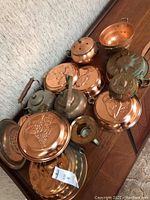 Photo showing a variety of copper and brass kitchen pieces including embossed copper moulds, a colander, kettles, and candle holders arranged on a wooden surface.