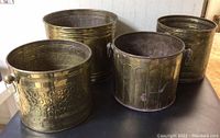 Four large round planters made of copper or brass with visible wear and patina, arranged on a black surface