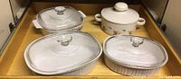 Four CorningWare covered dishes displayed side by side on wooden surface. Two oval dishes with glass lids, one square floral patterned dish with glass lid, and one round floral patterned dish with solid white lid.