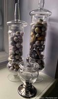 Photo of three decorative glass containers on a window ledge; two tall cylindrical jars filled with decorative balls and natural fillers, one smaller rounded jar with a silver base.