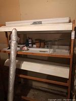 Full view of wooden grow rack with two grow lights, plastic trays on shelves, and accessories on middle shelf, roll of plastic sheeting leaning on side.