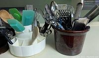 White plastic rotating holder with multiple silicone spatulas and wooden spoons on left, brown ceramic crock on right that holds metal kitchen utensils including graters, ladle, whisk, and others seen behind the holder.