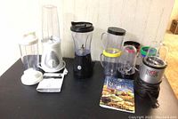 Overall lot showing three blenders with their cups, lids, extra blade, and recipe book on black surface against wall.