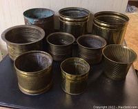 Full view showing all 9 metal pots arranged on dark surface, majority brass with one copper pot present, showing varied sizes and textures.