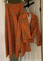 Three-piece Anne Klein outfit including a jacket, palazzo pants, and camisole in a retro floral geometric print, hanging on hangers against a white door.