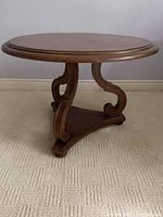 Full side view of solid wood round side table showing carved curved legs and triangular base platform on carpet.