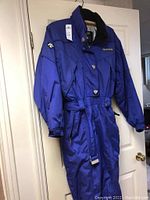 Front view of purple Descente ski suit hanging on door showing button and zipper closure, belt, pockets, and adjustable cuffs.