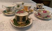 Seven vintage porcelain teacups with matching saucers arranged on a white tablecloth showing various floral and gold detailed designs.