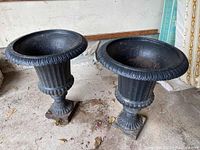 Photo of two black metal urns showing their full height and design from front, sitting on concrete.